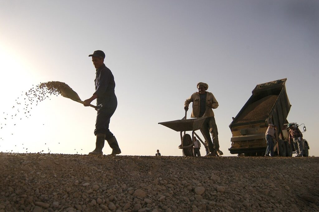 Trabalhadores espalhando cascalho em estrada rural com caminhão de entrega ao fundo