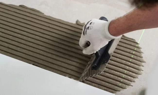 Mão com luva branca aplicando argamassa colante com desempenadeira dentada sobre superfície